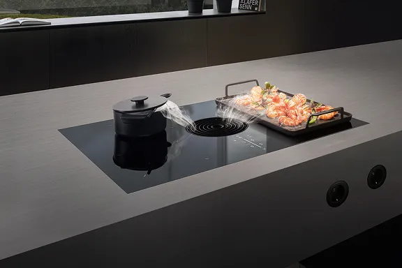 Detailaufnahme: Schwarzes BORA Induktionskochfeld mit integriertem Abzug, eingelassen in dunkle Arbeitsplatte. Dampf wird beim Braten von Garnelen abgesaugt.